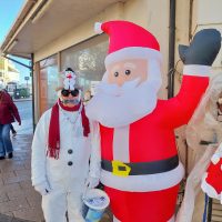 The photo features a person dressed as a snowman, complete with a white outfit, black buttons, and a red scarf. They are wearing a festive hat adorned with a snowman decoration and sunglasses, holding a charity collection bucket. Standing beside them is a large inflatable Santa Claus figure, waving cheerfully. The setting appears to be an outdoor area with a shopfront in the background, capturing a lighthearted and festive atmosphere.