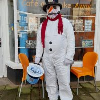 The photo shows a person dressed as a cheerful snowman, wearing a white outfit with black buttons, a red scarf, and a black top hat decorated with a snowman and festive details. They are holding a charity collection bucket marked with a blue label and QR code, as well as a walking cane. Behind them is a café window with the name "Relish Café and Take-Away" prominently displayed, along with colorful posters and signs. The setting creates a warm and welcoming atmosphere for their festive fundraising efforts.
