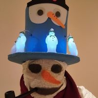 A person wears a snowman costume and a large, elaborate snowman hat. The hat is blue with a black top hat that reads "FROSTY" backward. It features googly eyes, a carrot nose, and three small, light-up snowman figures. The person's face is covered with a white cloth, decorated with a simple snowman face: black coal eyes, an orange carrot nose, and a black smile. A dark red scarf is wrapped around their neck, and they hold a black pipe in their mouth. The background is a plain, light tan wall.