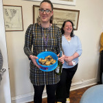 Two women stand in a room with framed pictures on the wall. The woman in the foreground wears a black and gold plaid dress, black tights, and dark shoes. She holds a blue plastic container of cookies and a bottle of white wine. A purple lanyard hangs around her neck. The woman behind her wears a light blue sweater, black pants, and yellow sneakers. She's smiling and holds a cell phone in her left hand. The room has parquet wood flooring. A partially visible doorway is on the left, and the edge of a light blue chair is visible on the right.