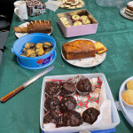 This image features a table set up for a gathering or party, covered with a green tablecloth and a variety of baked goods. In the foreground, there is a silver platter containing small cupcakes with pink and white frosting and gingerbread figures decorated with colorful candy. Surrounding the centerpiece are other desserts, including a loaf cake on a floral plate, cookies on a white tray, a pink box filled with frosted cupcakes, a tin of chocolates, and a plate of white chocolate discs with red sprinkles. The scene conveys a festive and welcoming atmosphere.