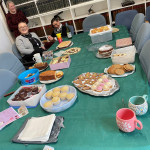 This image features a table set up for a gathering or party, covered with a green tablecloth and a variety of baked goods. In the foreground, there is a silver platter containing small cupcakes with pink and white frosting and gingerbread figures decorated with colorful candy. Surrounding the centerpiece are other desserts, including a loaf cake on a floral plate, cookies on a white tray, a pink box filled with frosted cupcakes, a tin of chocolates, and a plate of white chocolate discs with red sprinkles. The scene conveys a festive and welcoming atmosphere.