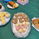This image features a table set up for a gathering or party, covered with a green tablecloth and a variety of baked goods. In the foreground, there is a silver platter containing small cupcakes with pink and white frosting and gingerbread figures decorated with colorful candy. Surrounding the centerpiece are other desserts, including a loaf cake on a floral plate, cookies on a white tray, a pink box filled with frosted cupcakes, a tin of chocolates, and a plate of white chocolate discs with red sprinkles. The scene conveys a festive and welcoming atmosphere.